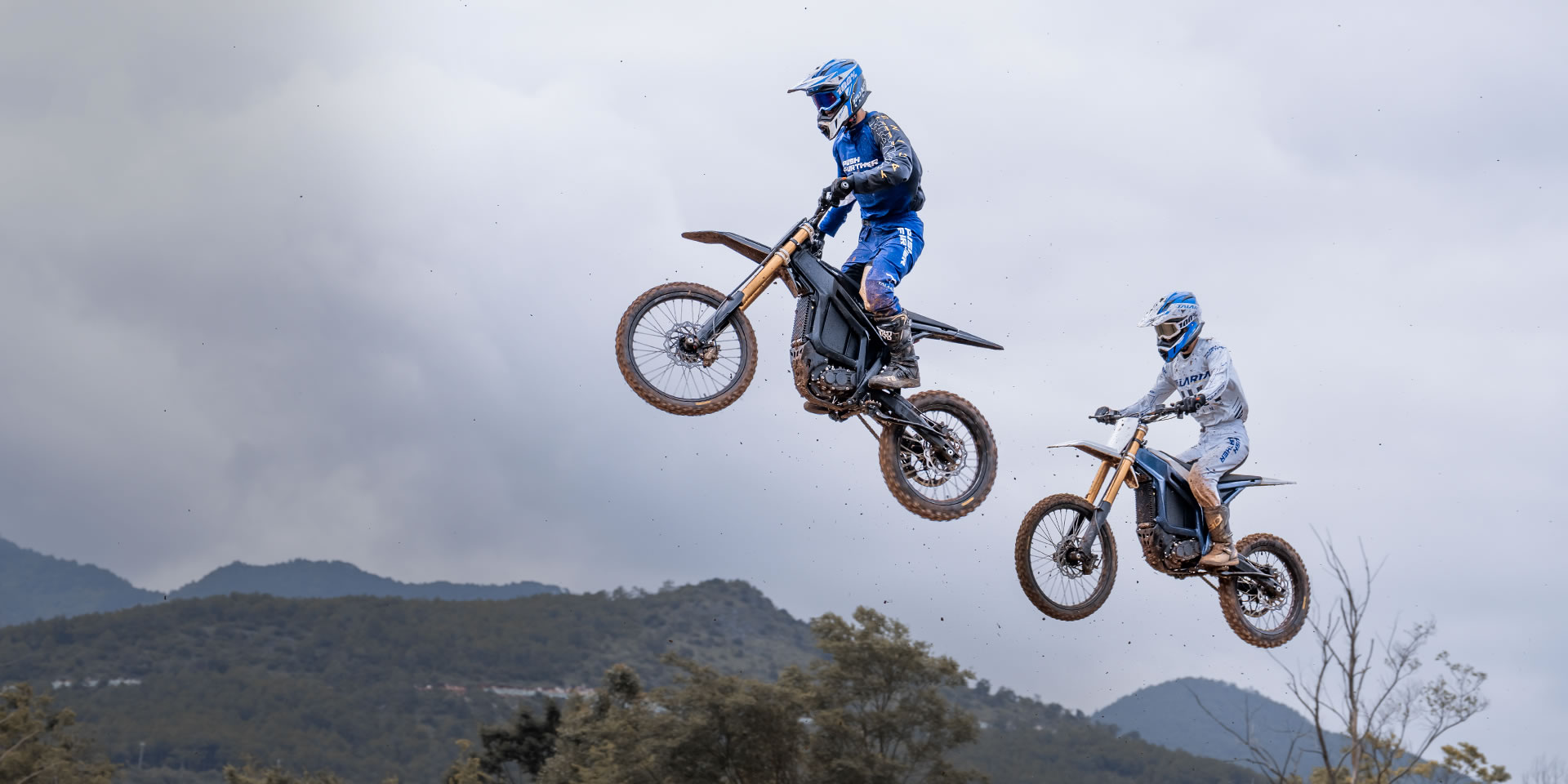 Two Talaria riders airborne above a dirt track, with mountains behind them