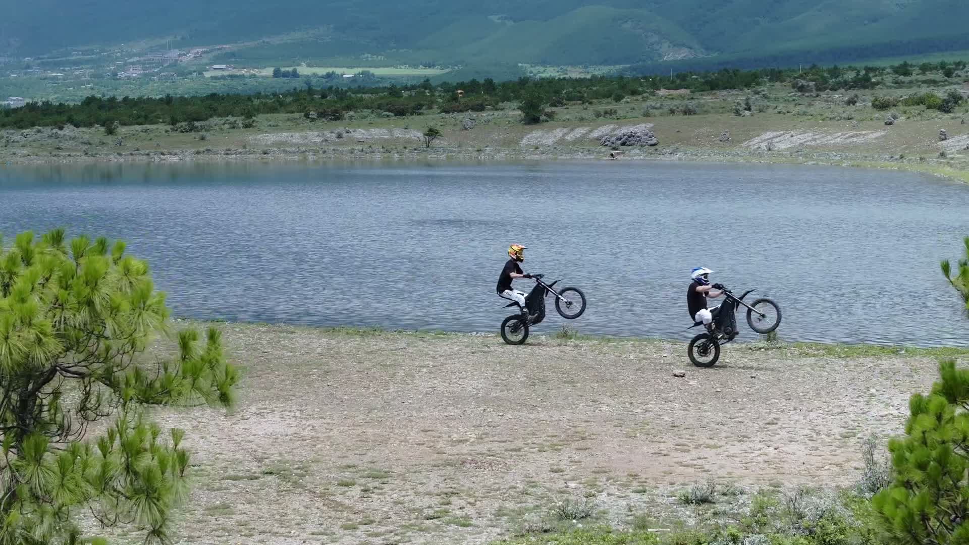 Two riders on Talaria motorcycles doing wheelies beside a lake